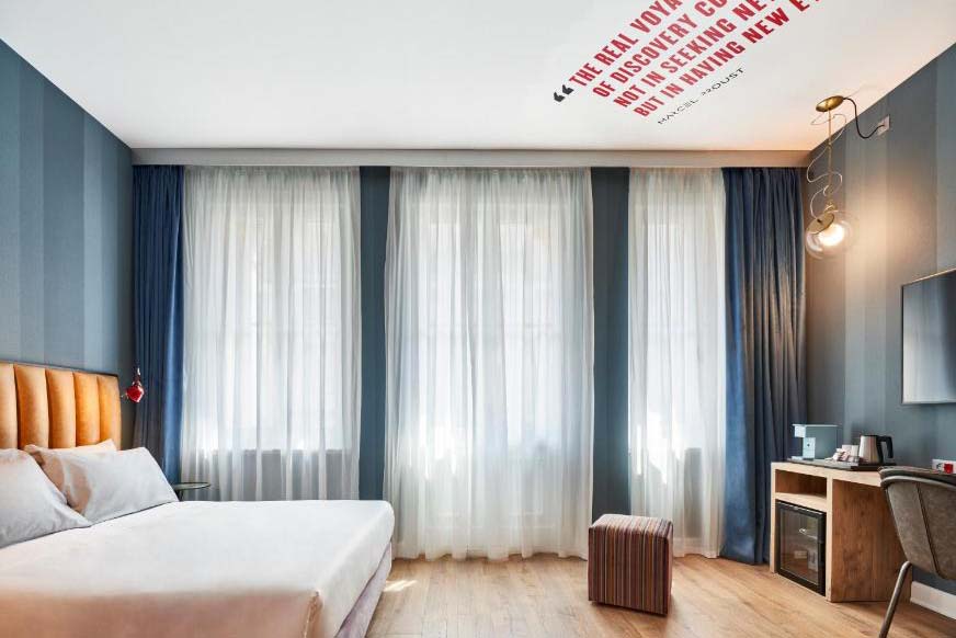 Helles Hotelzimmer mit Doppelbett, bodentiefen Fenstern, modernen Vorhängen, Holzboden und minimalistischer Einrichtung in ruhigem Design.