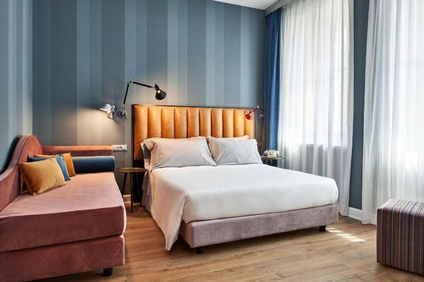 Modernes Hotelzimmer mit Doppelbett, gepolstertem Kopfteil, Schlafsofa, Holzfußboden und blau gestreiften Wänden bei Tageslicht.