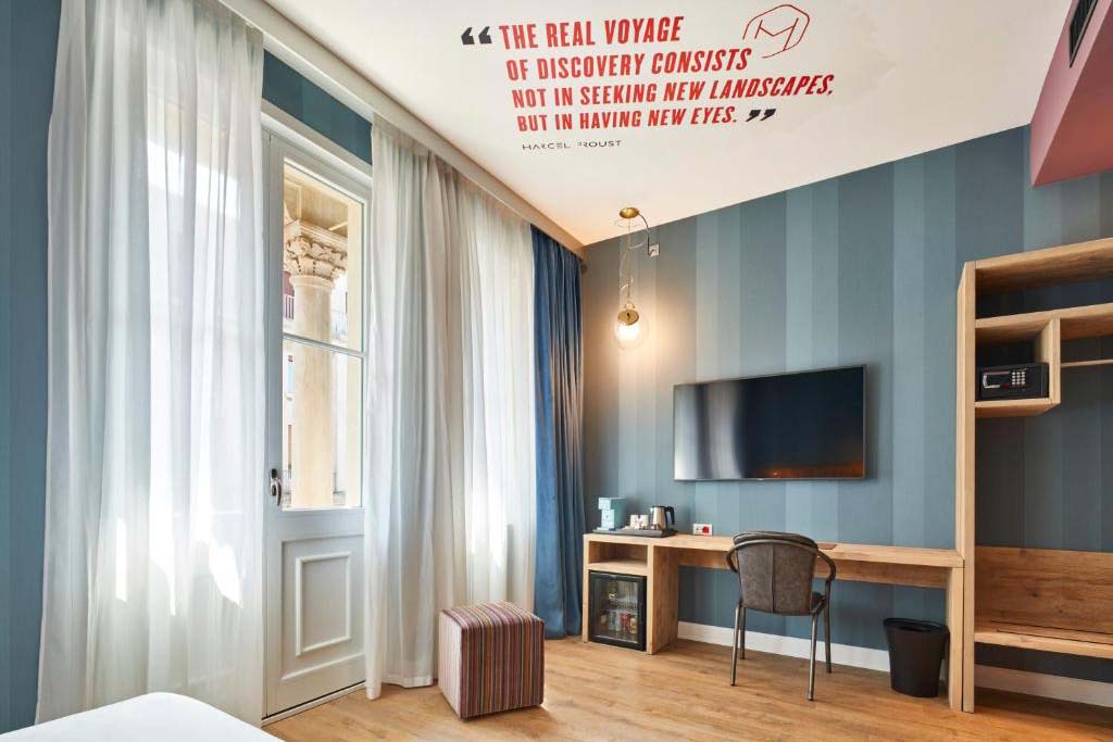 Helles Design-Hotelzimmer mit Schreibtisch, Fernseher, bodentiefen Vorhängen und Balkon­tür.