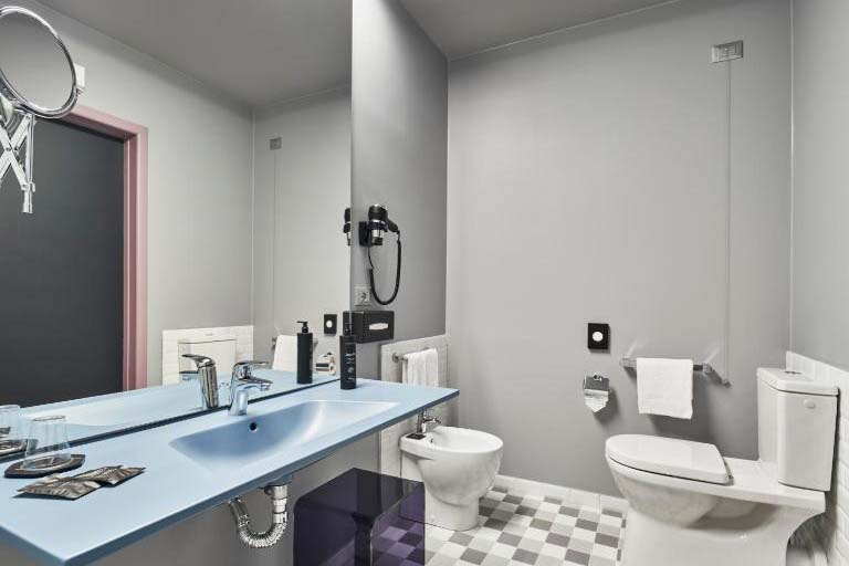 Modernes Hotelbad mit Waschbecken, Spiegel, WC, Bidet und grau-weiß gefliestem Boden in minimalistischem Design.