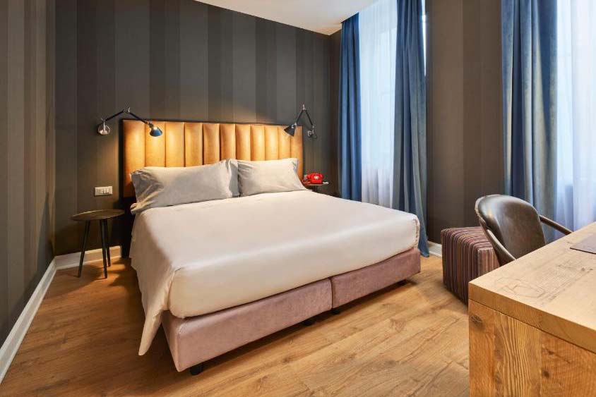 Modernes Hotelzimmer mit Doppelbett, gepolstertem Kopfteil, Holzfußboden, Schreibtisch und großen Fenstern mit Tageslicht.