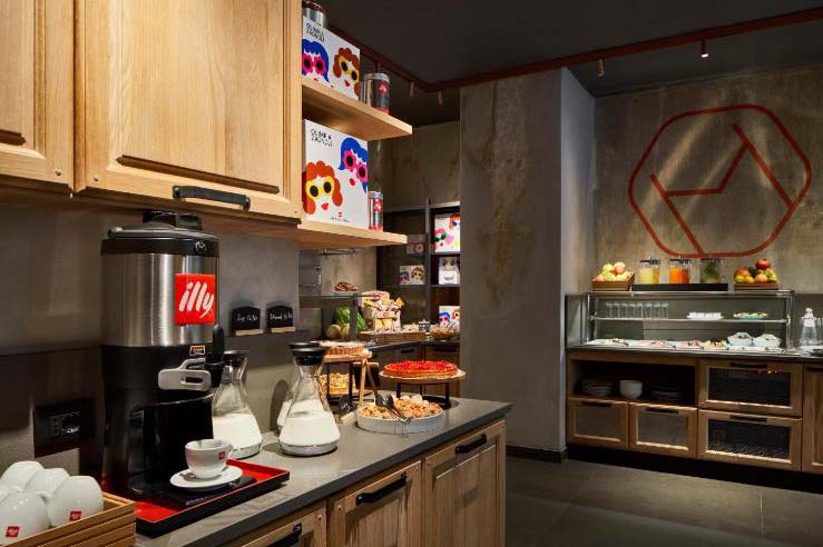 Stilvoller Frühstücksbereich im The Modernist Hotel mit Kaffeemaschine, Holzregalen, frischen Backwaren und moderner Industrial-Gestaltung.