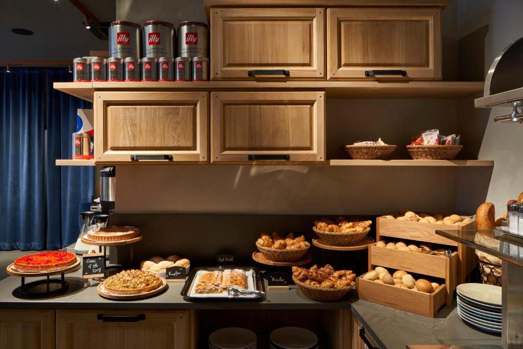 Frühstücksbuffet mit frischem Brot, Croissants, Kuchen und Kaffee in einer modern eingerichteten Hotelküche mit Holzschränken und warmer Beleuchtung.
