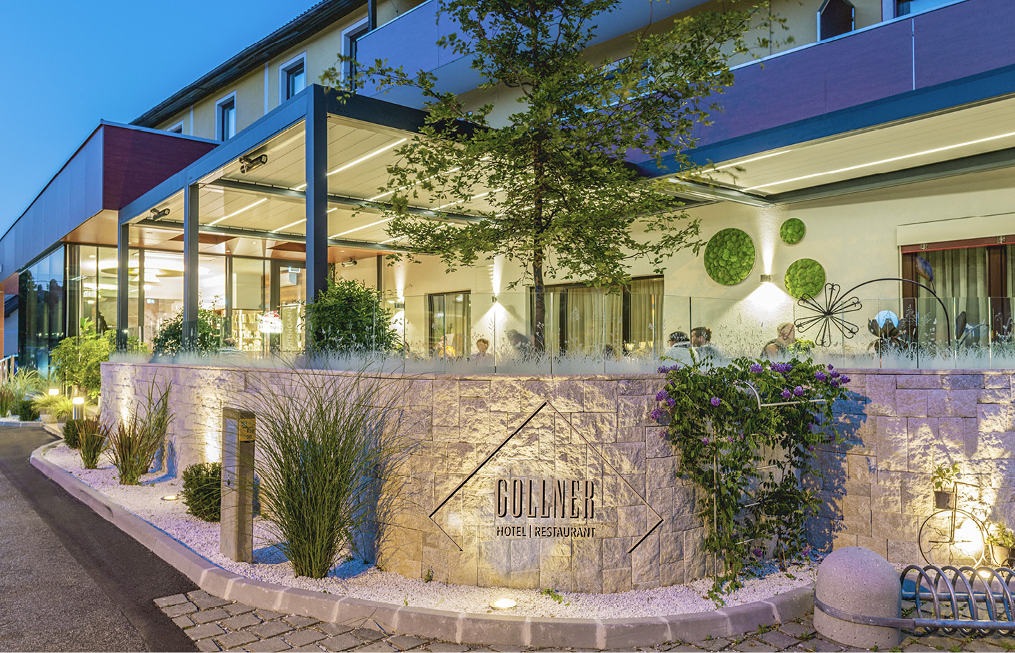 Außenansicht eines modernen Hotels mit beleuchtetem Eingangsbereich, Glasüberdachung und bepflanztem Vorgarten mit Steinmauer und Schriftzug