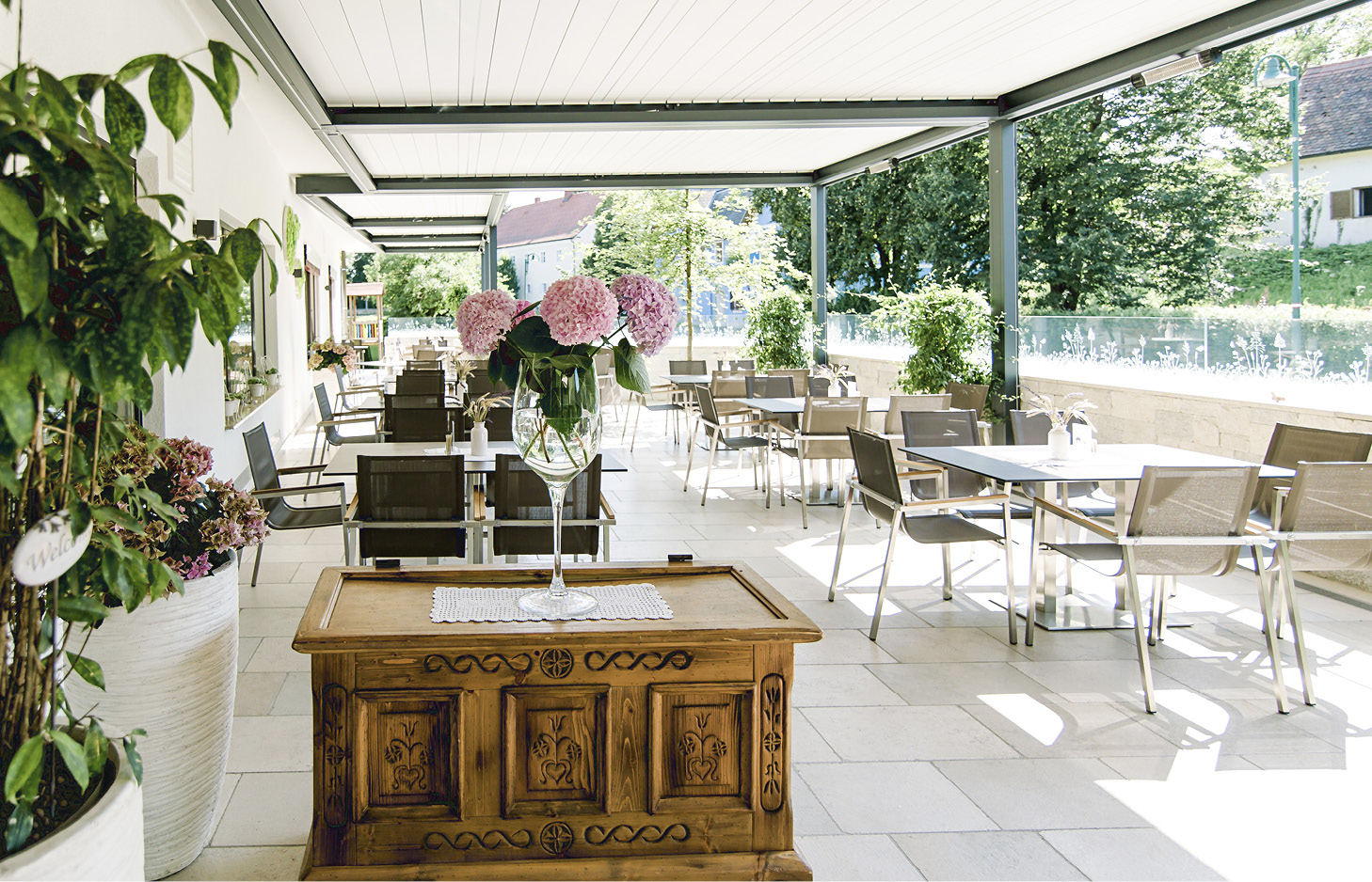 Überdachte Terrasse mit Tischen und Stühlen sowie einer Holzkommode mit Vase und Blumen im Vordergrund