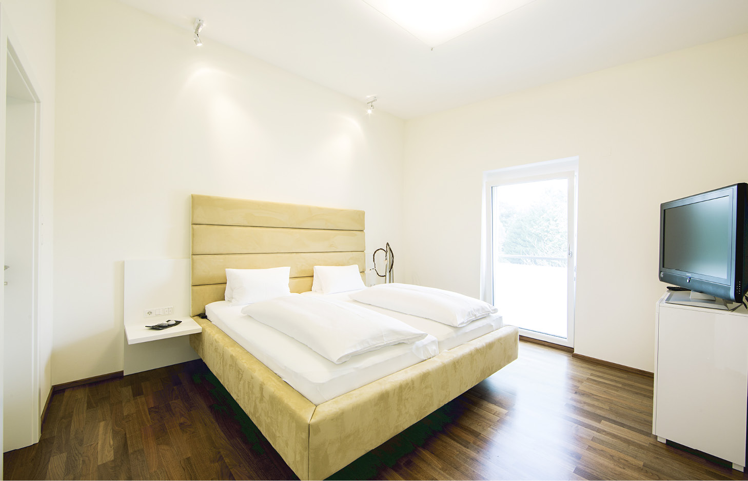 Hotelzimmer mit Doppelbett, zwei weißen Kissen, Nachttisch, Stehlampe, Fernseher auf Kommode und Fenster mit Blick nach draußen