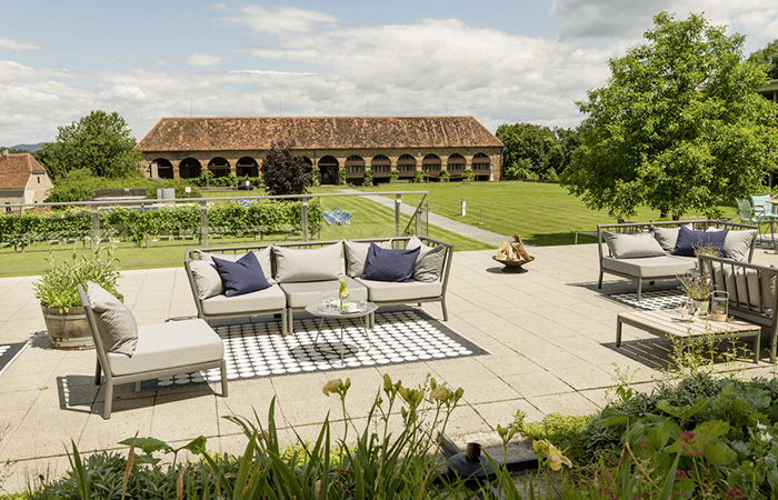 Terrasse mit moderner Sitzgruppe aus Polstermöbeln und Blick auf eine historische, langgestreckte Backsteinhalle mit Rundbögen