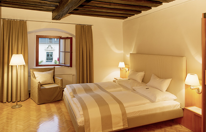 Hotelzimmer mit Doppelbett, Sessel vor offenem Fenster und warmem Licht von Steh- und Wandlampen