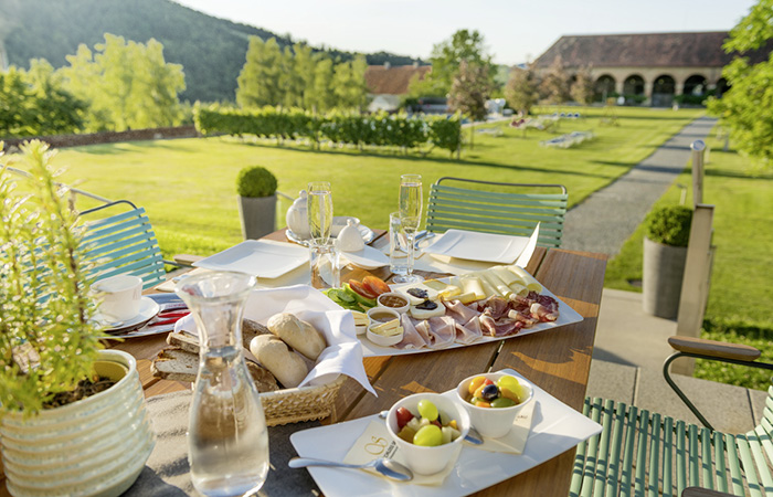 Tisch auf Terrasse mit gedecktem Frühstück, darunter Obstschalen, Aufschnitt, Brot und Getränke, im Hintergrund Garten und Schlossgebäude