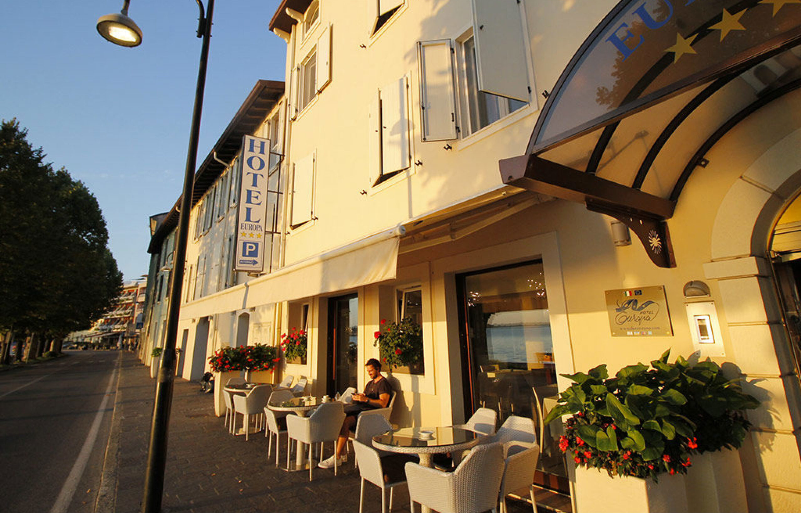 Straßencafé mit Tischen und Stühlen vor einem hellen Hotelgebäude bei Sonnenuntergang