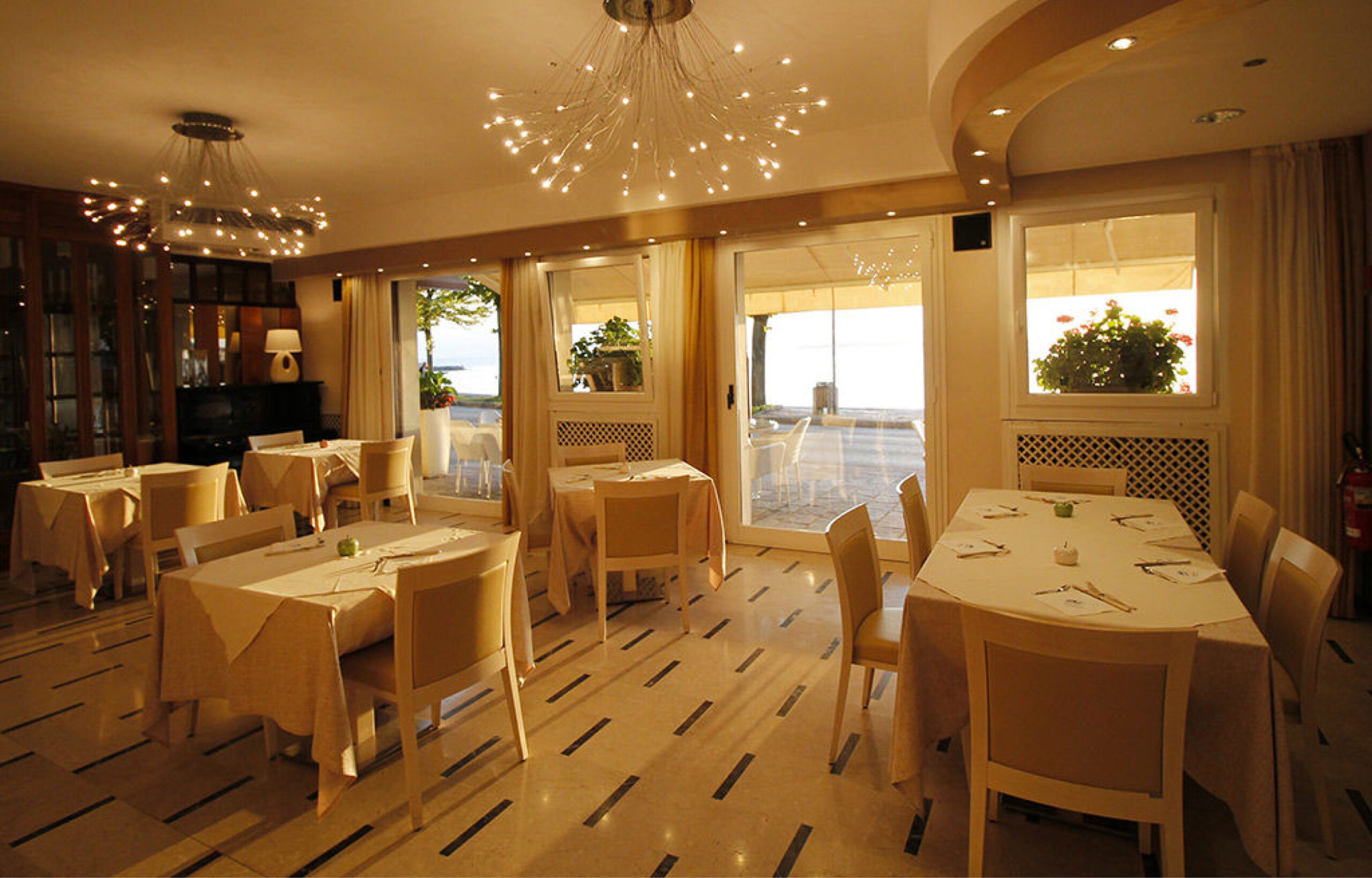 Innenraum eines Restaurants mit gedeckten Tischen und großen Fenstern mit Blick auf eine Terrasse und das Meer