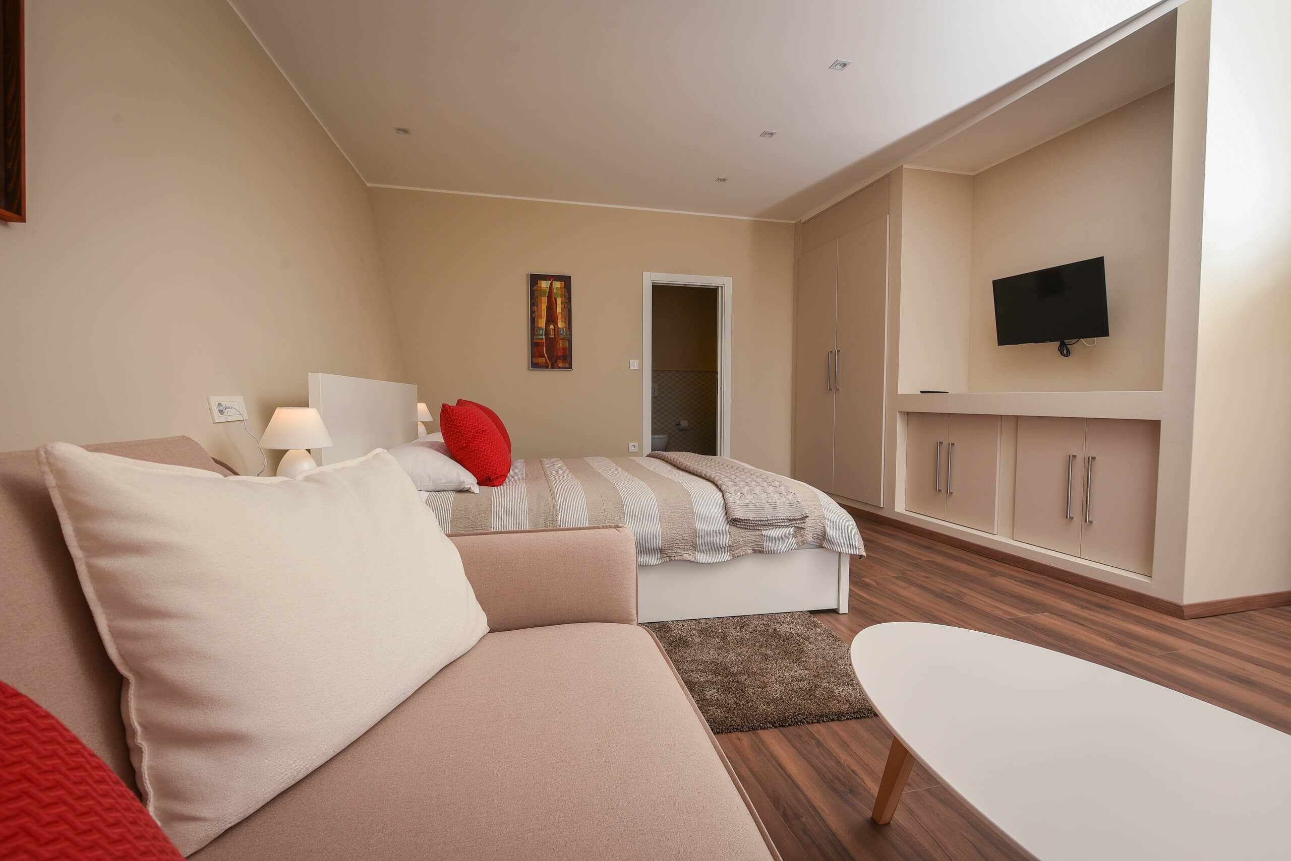 Hotelzimmer mit Doppelbett, Sofa mit Kissen, Wandbild, Wandfernseher und eingebautem Schrank