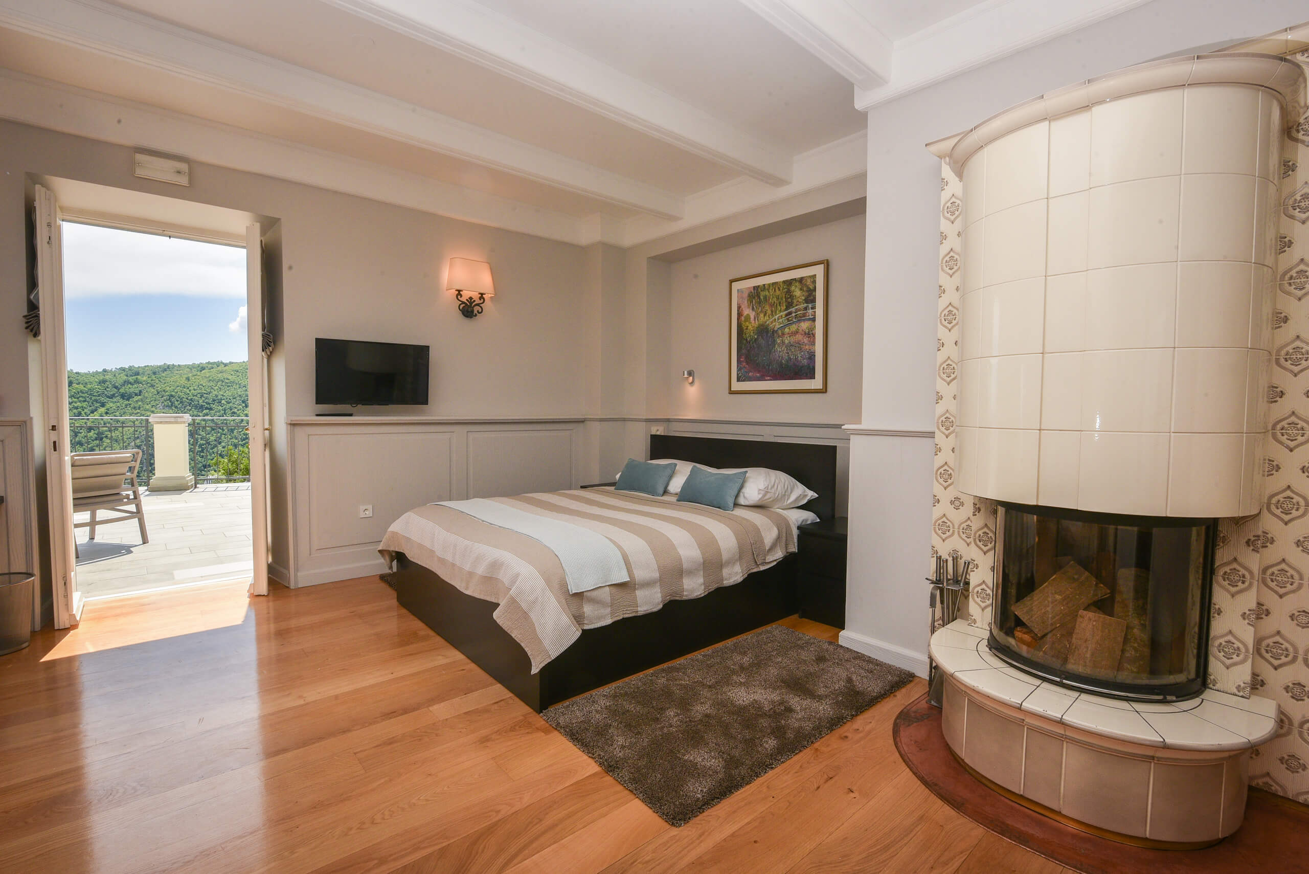Schlafzimmer mit Doppelbett, Kamin mit Kacheln, Holzboden und offener Tür zu Terrasse mit Blick auf bewaldete Hügel