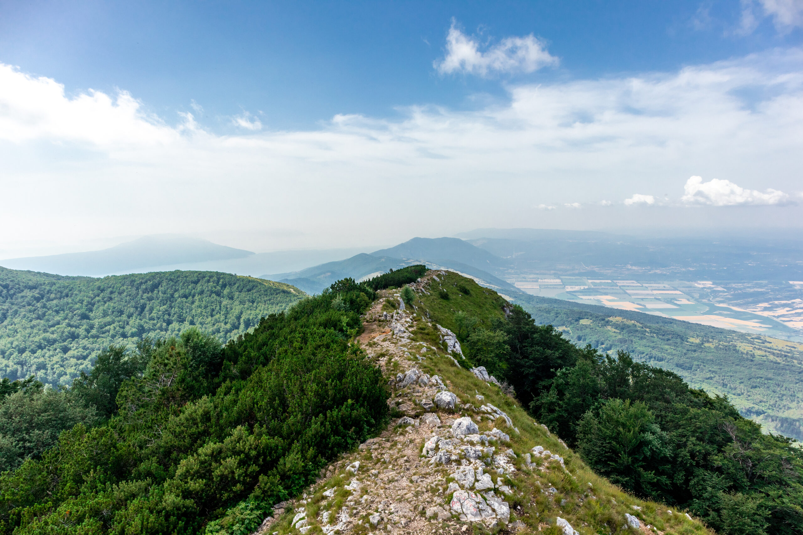 Bergkamm mit felsigem Pfad und bewaldeten Hängen unter blauem Himmel mit Wolken